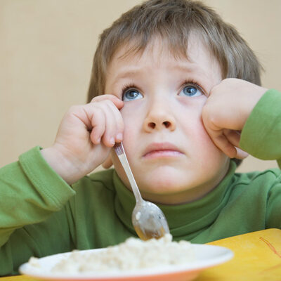 A frowning young boy is looking up from his bowl of food
